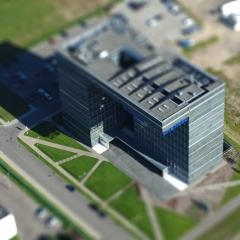 Tumša stikla biroju ēka starp zālieniem un gājēju celiņiem, skatīta no augšas.