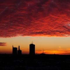 Spilgti sarkani mākoņi virs tumša pilsētas silueta un augstceltnēm.
